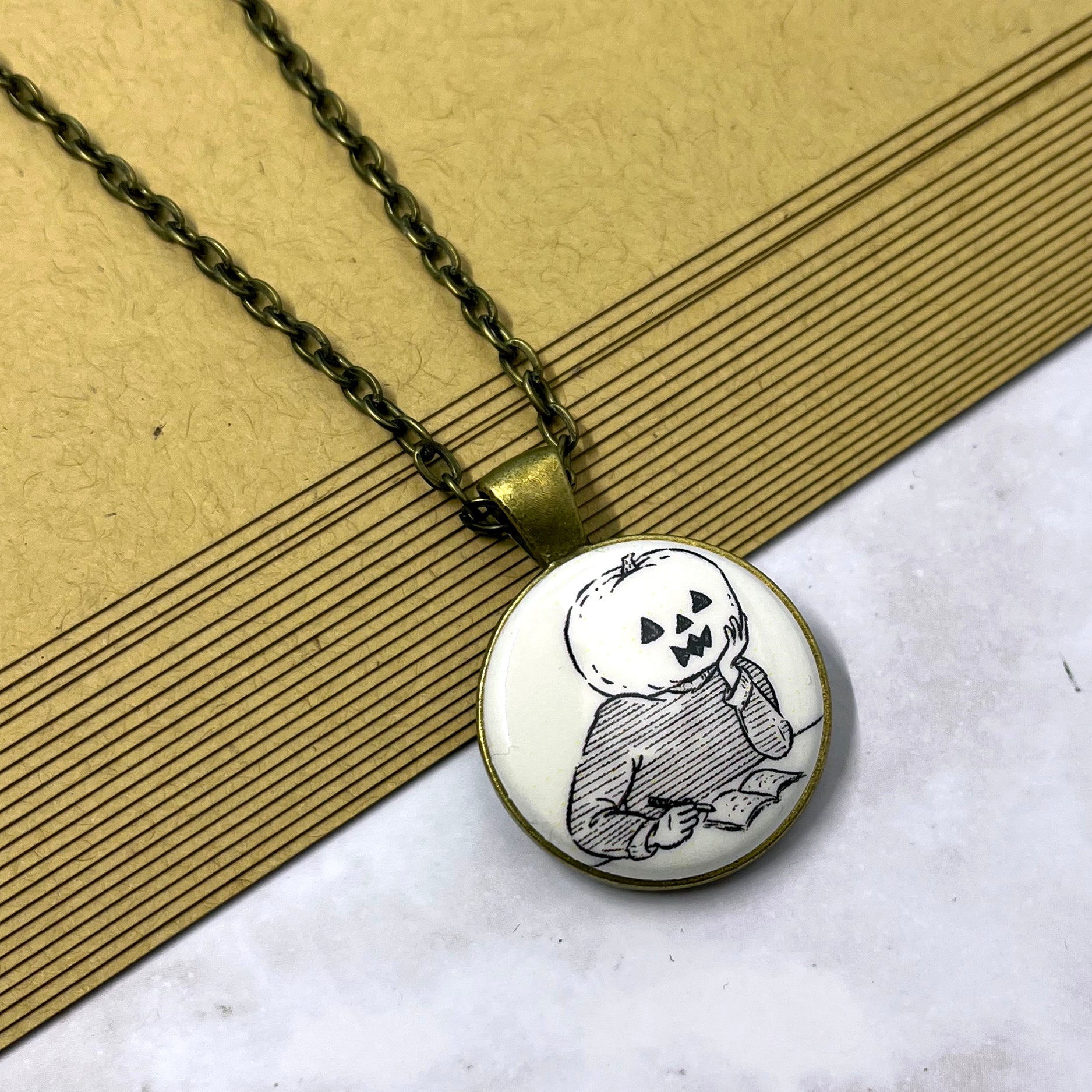 Photo shows a necklace on a stack of brown paper. The pendant is an illustrated line drawing of Pumpkinhead. Pumpkinhead is at a desk or table and appears to have paused while writing in a notebook and caught in mid-thought. His elbow leans on the table and his chin rests in his hand. He wears a striped sweater. The pendant is a cream white color on a metal backing. The chain and pendant backing are antique bronze in color.