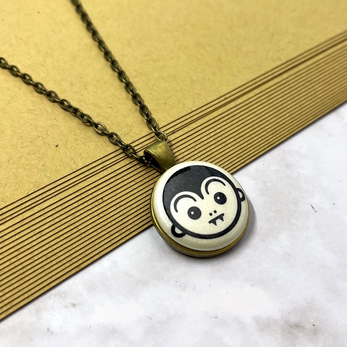 Photo shows a necklace over a stack of brown paper. The pendant shows a black and white illustrated graphic of a vampire with a widow’s peak and fangs. The white background on the pendant is a shimmery cream white. The metal chain is an antique bronze in color.